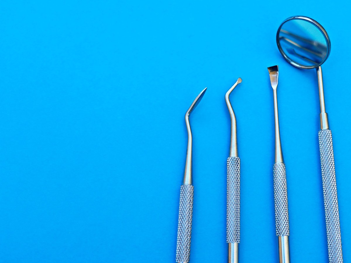 Four stainless steel dental instruments, including a mirror and a scaler, laid on a bright blue surface