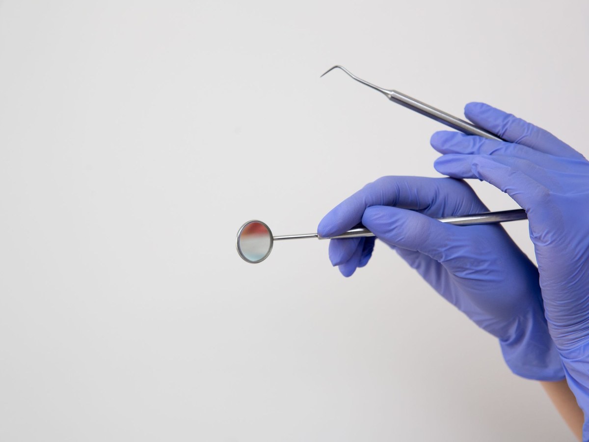 Blue-gloved hands holding a dental mirror and a probe against a white background